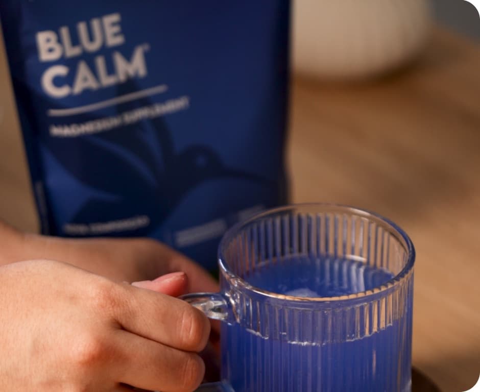 Mão segurando uma caneca de vidro com bebida azul ao lado do sachê Blue Calm Mão segurando uma caneca de vidro com bebida azul ao lado do sachê Blue Calm